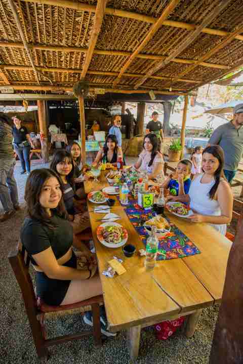 Grupo de amigos comiendo en una mesa larga bajo la palapa